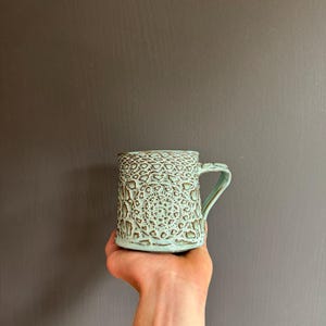 Könnte beinhalten: Ein Keramikbecher mit hellblauer Glasur und einem dunkelbraunen, strukturierten Reliefmuster aus komplexen geometrischen Formen und kreisförmigen Motiven. Der Becher hat einen stabilen Henkel und einen zylindrischen Körper.