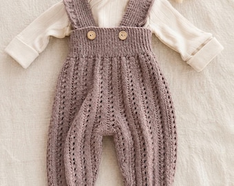 May include: A mauve knitted overall with a textured vertical stitch pattern, featuring two small wooden buttons at the waist and ruffled shoulder straps, layered over a long-sleeved cream-colored ribbed shirt with a crossover neckline.
