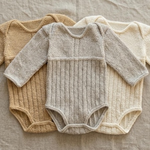 May include: Three knitted baby bodysuits in neutral colors: tan, gray, and cream. Each has long sleeves and a ribbed texture on the front. The bodysuits are laid out on a neutral-colored fabric.