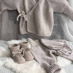 May include: A beige knitted baby set consisting of a long-sleeved wrap cardigan with a side tie closure, a pair of matching booties with ribbon ties, a pair of ribbed-cuff leggings, and a matching bonnet with long ties, all in a textured knit.
