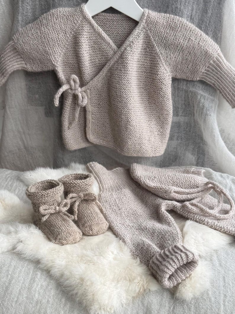 May include: A beige knitted baby set consisting of a long-sleeved wrap cardigan with a side tie closure, a pair of matching booties with ribbon ties, a pair of ribbed-cuff leggings, and a matching bonnet with long ties, all in a textured knit.