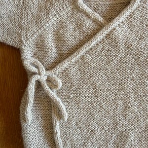 May include: A beige long-sleeved wrap cardigan featuring a textured knit pattern. The garment includes a V-neckline and a side tie closure with matching knitted strings that form a bow. The knit construction is consistent throughout the piece.