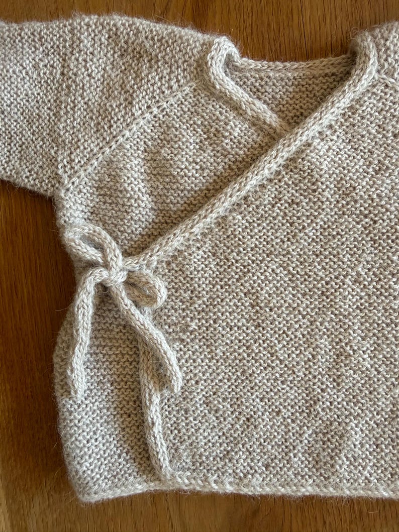 May include: A beige long-sleeved wrap cardigan featuring a textured knit pattern. The garment includes a V-neckline and a side tie closure with matching knitted strings that form a bow. The knit construction is consistent throughout the piece.