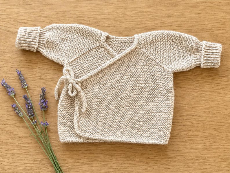 May include: A beige long-sleeved wrap cardigan made of knit fabric with ribbed cuffs and a side tie closure. The garment features a V-neckline and a simple, solid-colored design. Several sprigs of purple lavender are positioned to the left.