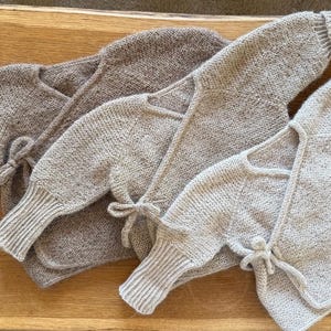 May include: Three long-sleeved wrap-style cardigans in varying shades of beige, tan, and light brown. Each garment features a knitted texture, ribbed cuffs at the wrists, and a side-tie closure with matching knitted strings to secure the front overlap.