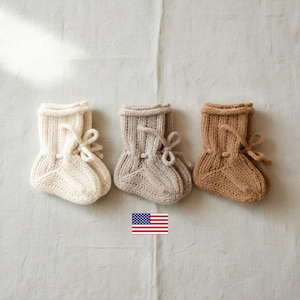 May include: Three pairs of knitted booties in cream, light beige, and tan, each featuring a ribbed cuff and a decorative bow tie detail on the front. A small rectangular icon of the United States flag is positioned below the center bootie.