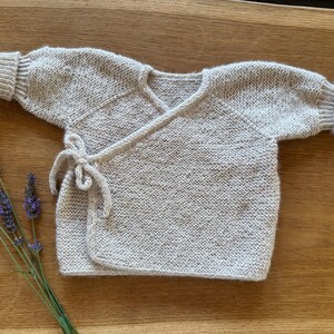 May include: A long-sleeved wrap-style cardigan knitted in a light beige yarn with subtle brown flecks. The garment features a V-neckline, ribbed cuffs at the wrists, and a side tie closure made from matching yarn. The texture is a consistent garter stitch.