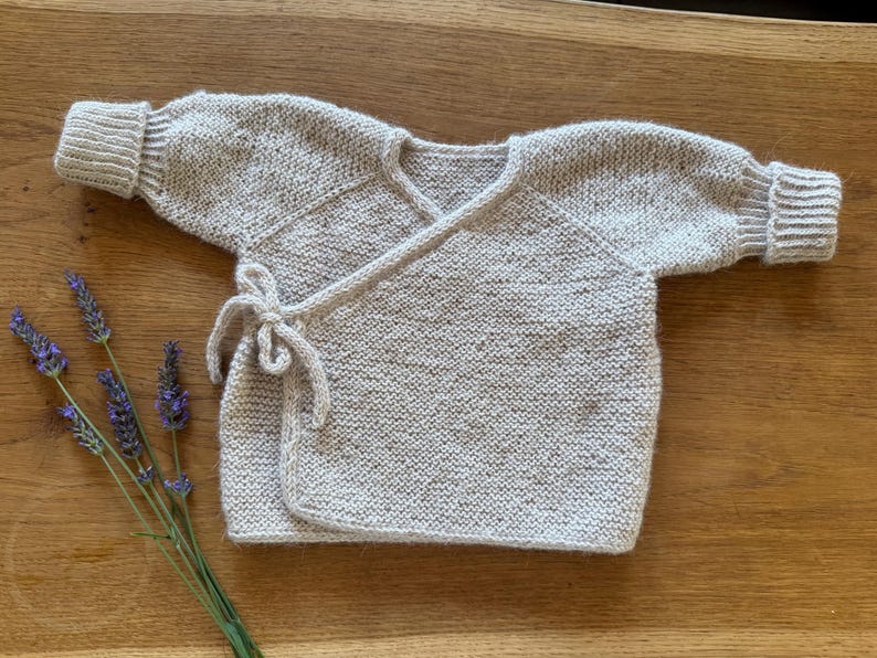 May include: A long-sleeved wrap-style cardigan knitted in a light beige yarn with subtle brown flecks. The garment features a V-neckline, ribbed cuffs at the wrists, and a side tie closure made from matching yarn. The texture is a consistent garter stitch.