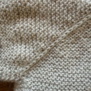 May include: A beige knitted garment featuring a garter stitch pattern with a visible diagonal seam connecting two sections of the fabric. The texture appears soft and slightly fuzzy, with a consistent light tan color throughout the entire piece.
