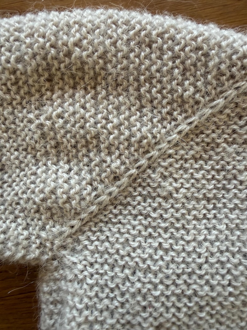 May include: A beige knitted garment featuring a garter stitch pattern with a visible diagonal seam connecting two sections of the fabric. The texture appears soft and slightly fuzzy, with a consistent light tan color throughout the entire piece.