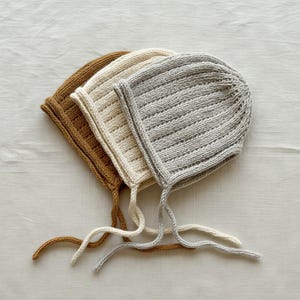 May include: Three knitted bonnets with ribbed texture and chin ties, arranged in a stack. The bonnets are colored in shades of mustard yellow, cream, and light gray. Each bonnet features a rounded shape with a structured edge and long ties.
