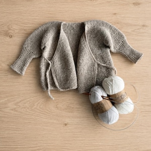 May include: A beige knit wrap cardigan with long sleeves and ribbed cuffs, featuring a tie closure at the front. Two balls of white yarn with labels reading Alpaca and a circular knitting needle are positioned next to the garment.