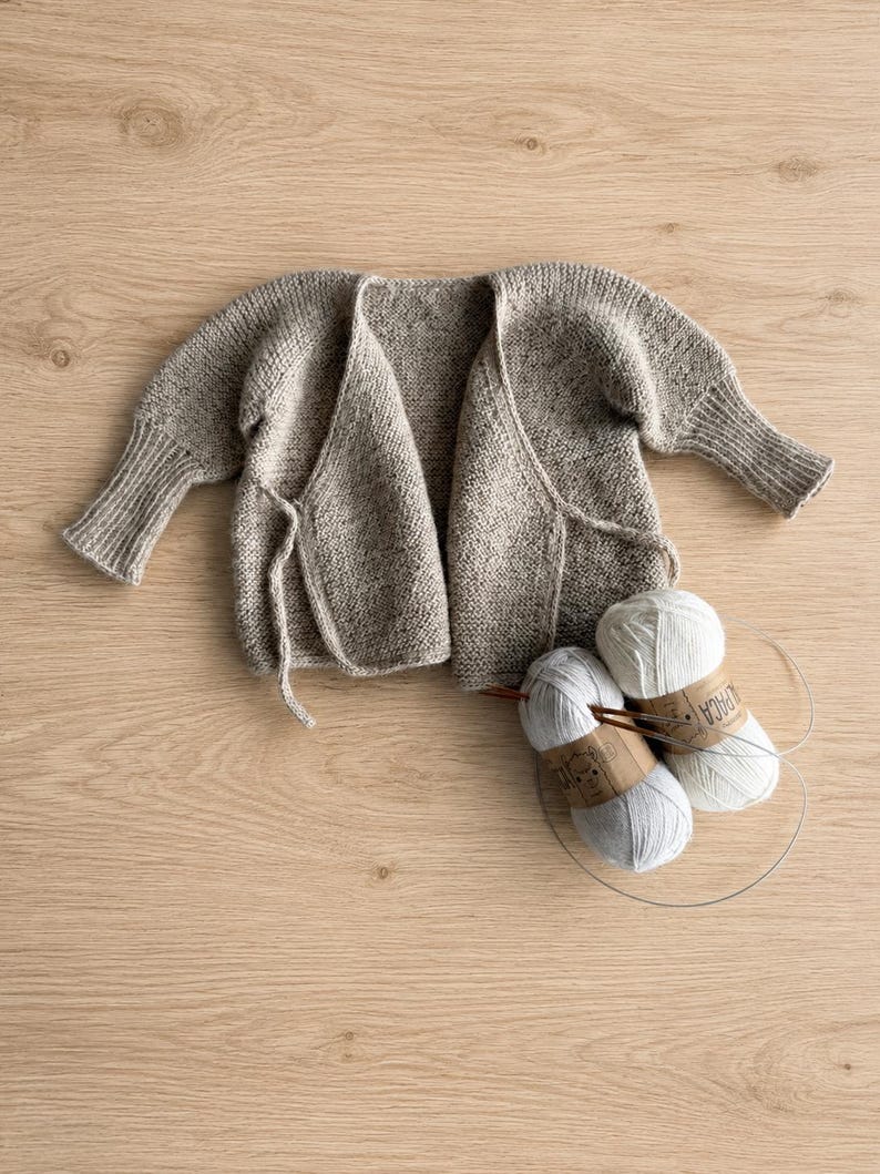 May include: A beige knit wrap cardigan with long sleeves and ribbed cuffs, featuring a tie closure at the front. Two balls of white yarn with labels reading Alpaca and a circular knitting needle are positioned next to the garment.