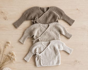 May include: Three long-sleeved wrap-style knit sweaters for infants, arranged vertically in a gradient from dark brown to light beige. Each sweater features a side-tie closure with matching knit strings and ribbed cuffs on the sleeves.