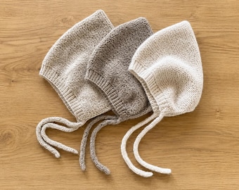 May include: Three knitted bonnets in light beige, medium brown, and cream colors. Each bonnet features a textured garter stitch pattern, a ribbed edge around the face opening, and two long knitted ties extending from the bottom corners.