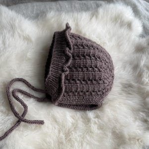 Peut inclure: Un bonnet tricoté mauve présentant un motif de points texturés, un bord côtelé avec une finition à volants et deux longs liens tricotés s'étendant sur les côtés. Le bonnet est fabriqué avec un fil doux et affiche une couleur uniforme.