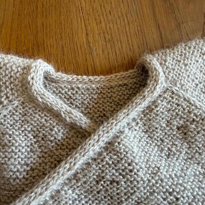 May include: A beige knitted garment featuring a wrap-style neckline with a thick, textured border. The piece is crafted from a soft, fuzzy yarn in a garter stitch pattern, showcasing a clean, overlapping design at the collar and raglan sleeves.