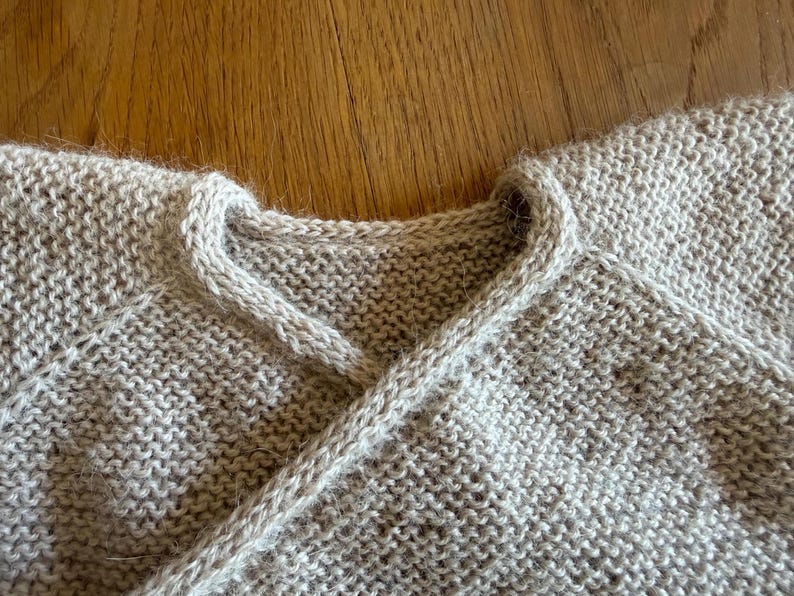 May include: A beige knitted garment featuring a wrap-style neckline with a thick, textured border. The piece is crafted from a soft, fuzzy yarn in a garter stitch pattern, showcasing a clean, overlapping design at the collar and raglan sleeves.