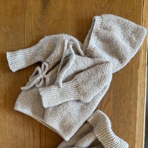 May include: A light beige knitted hooded sweater with long sleeves and ribbed cuffs. The garment features a wrap-around design that secures at the side with a matching knitted tie. The texture is consistent throughout, showcasing a simple knit pattern.