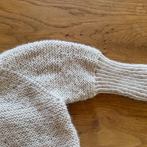 May include: A light beige knitted sweater featuring a textured garter stitch pattern throughout the body and sleeves. The garment includes long sleeves that transition into a ribbed cuff at the wrist, with a simple neckline and a soft, fuzzy finish.