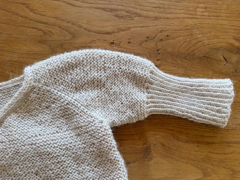 May include: A light beige knitted sweater featuring a textured garter stitch pattern throughout the body and sleeves. The garment includes long sleeves that transition into a ribbed cuff at the wrist, with a simple neckline and a soft, fuzzy finish.