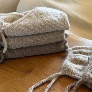 May include: A stack of three folded beige knit garments with tie closures, accompanied by two matching knit bonnets with long ties. The items feature a textured, speckled yarn construction in neutral earth tones, showcasing a consistent stitch pattern throughout the set.