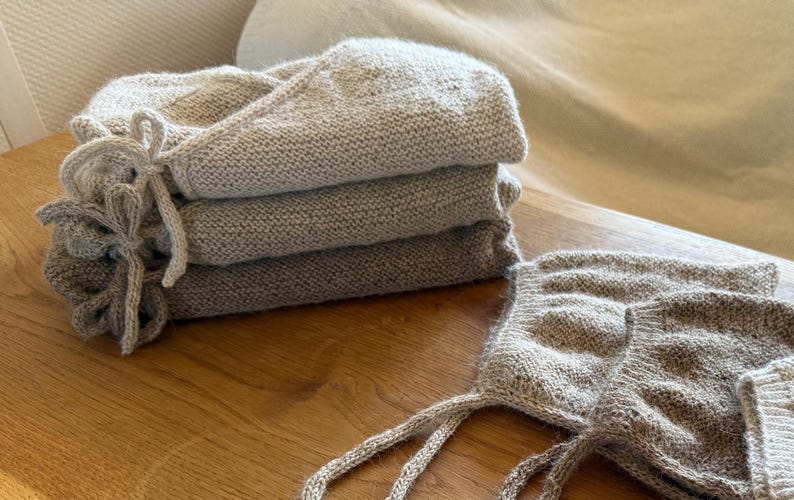 May include: A stack of three folded beige knit garments with tie closures, accompanied by two matching knit bonnets with long ties. The items feature a textured, speckled yarn construction in neutral earth tones, showcasing a consistent stitch pattern throughout the set.