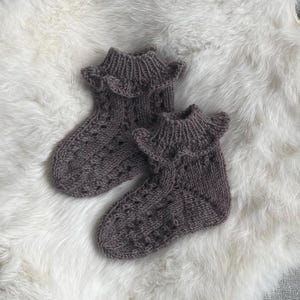 May include: A pair of ankle-length knitted socks in a muted taupe color. The socks feature a ribbed cuff with a decorative ruffled edge and a textured cable knit pattern running down the foot and ankle, crafted from a soft yarn material.