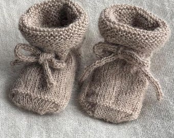 May include: A pair of light brown knitted booties featuring a garter stitch cuff and a matching tie closure at the front. The booties are crafted from a soft, textured yarn with a fuzzy appearance, showcasing a simple design with rounded toes.