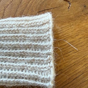 May include: A beige knitted cuff featuring a ribbed texture with a fuzzy, brushed surface. The fabric displays a consistent vertical stitch pattern with loose fibers extending from the edge, showcasing the soft, textured nature of the yarn.