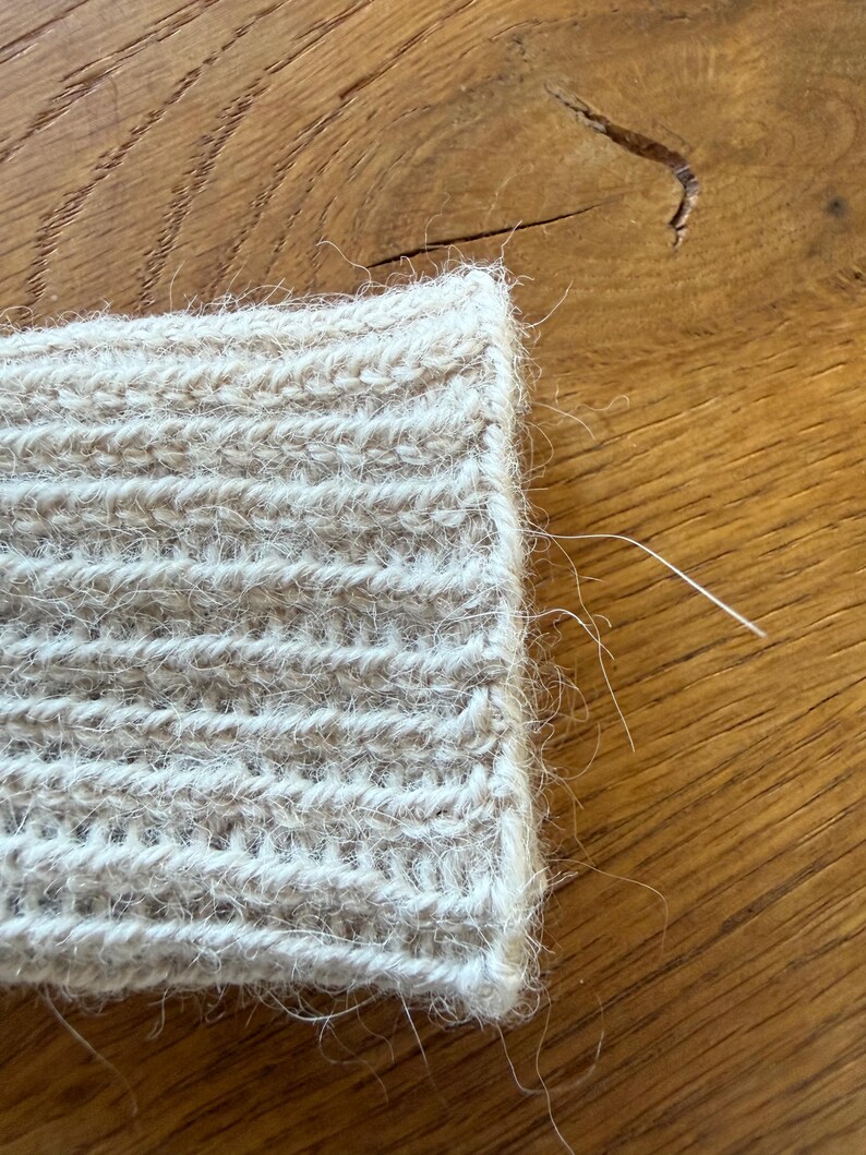 May include: A beige knitted cuff featuring a ribbed texture with a fuzzy, brushed surface. The fabric displays a consistent vertical stitch pattern with loose fibers extending from the edge, showcasing the soft, textured nature of the yarn.