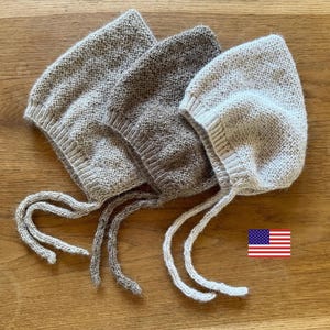 Può includere: Tre cuffiette lavorate a maglia in varie tonalità di beige, marrone chiaro e crema. Ogni cuffietta presenta un motivo a maglia testurizzato, un bordo a coste attorno all'apertura del viso e due lunghi lacci lavorati agli angoli.