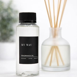 May include: A clear glass bottle with a black cap and a black label that reads "MY WAY FRAGRANCE OIL". Next to it is a clear glass diffuser bottle with wooden reeds. The fragrance oil includes Oud Wood, Leather, and Sandalwood.