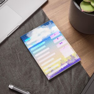 Puede incluir: Un bloc de notas con un diseño colorido con temática de cielo. El diseño incluye tonos azules, amarillos y morados, con líneas de cuadrícula e iconos. El bloc de notas está sobre una superficie de madera junto a un portátil y una planta.