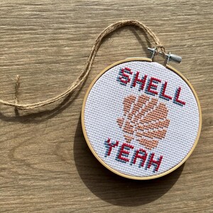 May include: A circular wooden embroidery hoop containing white fabric with a cross-stitch design of a peach-coloured seashell centred between the blue and red text that reads SHELL YEAH. A piece of brown twine is attached to the metal tension screw.