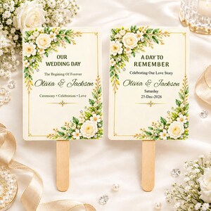 Op de afbeelding: Twee rechthoekige trouwwaaier met houten handvatten, voorzien van crèmekleurige achtergronden, gouden randen en bloemillustraties van witte rozen en groene bladeren. De tekst luidt: OUR WEDDING DAY, The Begining Of Forever, Olivia & Jackson, Ceremony, Celebration, Love, A DAY TO REMEMBER, Celebrating Our Love Story, Olivia & Jackson, Saturday, 25-Dec-2026.