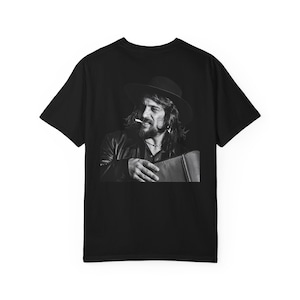 Può includere: T-shirt nero con un'immagine in bianco e nero di un uomo con cappello e giacca di pelle, che fuma una sigaretta. L'immagine è centrata sulla parte anteriore della maglietta e tiene una valigetta.