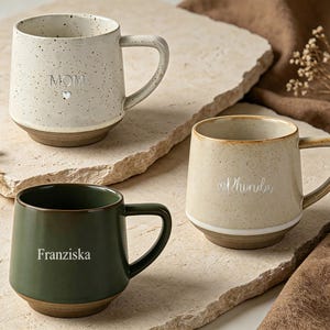 May include: Three ceramic mugs with handles, featuring a speckled off-white mug with the text MOM and a heart, a speckled beige mug with the text Rhonda and a heart, and a solid dark green mug with the text Franziska printed on the front.