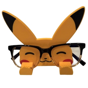 Pode incluir: Um suporte de óculos amarelo em forma de Pikachu com óculos de armação preta. O suporte tem orelhas com pontas pretas e um rosto sorridente com bochechas vermelhas. Os óculos são mantidos no lugar por pequenos suportes amarelos.