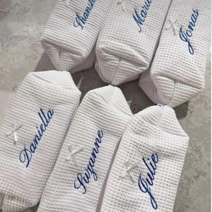 Könnte beinhalten: Sechs weiße Stoffbeutel mit Waffelstruktur, jeweils mit einer kleinen weißen Schleife und einem in blauer Schreibschrift gestickten Namen versehen. Die auf den Beuteln sichtbaren Namen sind Shanshan, Marius, Jonas, Daniella, Suzanne und Julie.