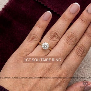 May include: A gold solitaire ring featuring a round-cut 1CT diamond set in a four-prong setting. The text 1CT SOLITAIRE RING is displayed on the image. The ring is worn on a finger, showcasing the clear, faceted stone and the polished gold band.