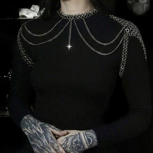 May include: Silver chainmail shoulder piece with a star pendant. The jewelry features multiple strands of delicate chains and ringmail, creating a layered effect. The piece is worn over a black long-sleeve top, with a dark background.