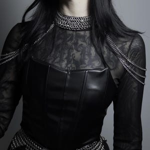 Puede incluir: Un corsé de cuero negro con detalles de cota de malla. El corsé se combina con una blusa negra transparente, un collar y un cinturón de cota de malla. La imagen tiene una estética gótica.