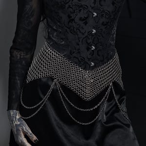 May include: A black corset top featuring a tonal damask pattern and silver-toned front clasps, paired with a silver chainmail waist belt that has draped chain accents hanging over a smooth black skirt. The ensemble is worn with a sheer black long-sleeve top.
