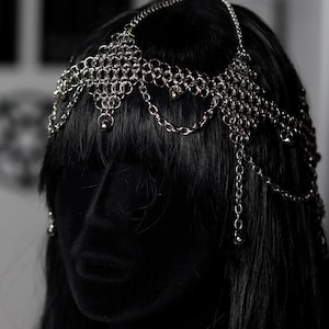 May include: A silver-toned chainmail headpiece featuring intricate interlocking metal rings and delicate hanging chains. The headpiece is constructed with a woven mesh pattern that drapes across the forehead and sides, accented by small metallic beads.