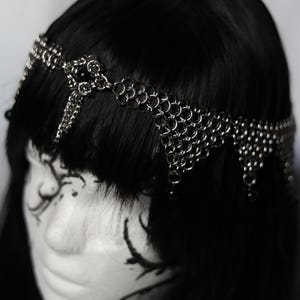 May include: A silver chainmail headpiece with a draped design. The headpiece is made of small, interconnected metal rings and is worn on black hair. The headpiece has a delicate, intricate appearance.