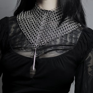 Puede incluir: Un collar de cota de malla plateada con un colgante de espada. El collar está hecho de pequeños anillos de metal entrelazados y se coloca alrededor del cuello. El colgante es una pequeña espada plateada. La persona lleva una camiseta negra.