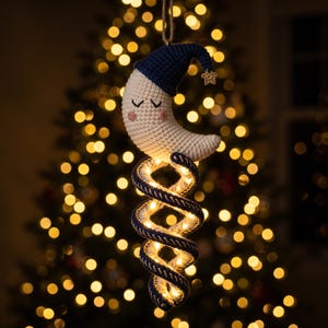 May include: A crocheted crescent moon ornament with a dark blue nightcap and a small gold star charm. Below the moon hangs a spiral tail made of dark blue and white rope, illuminated by integrated warm white LED fairy lights on a jute string.