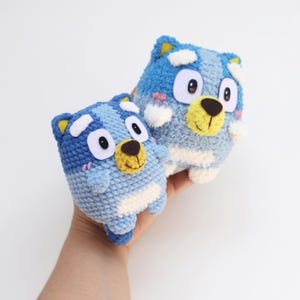 May include: Two handmade blue and white crochet toys with yellow and brown details. The toys have large black eyes and yellow ears. The toys are held in a person's hand.
