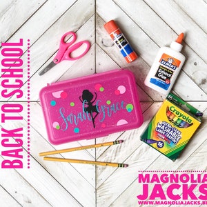 May include: Pink glitter pencil case with a personalized design that says "Sarah Grace" and a silhouette of a ballerina. The case is surrounded by school supplies including pencils, glue, crayons, and scissors.